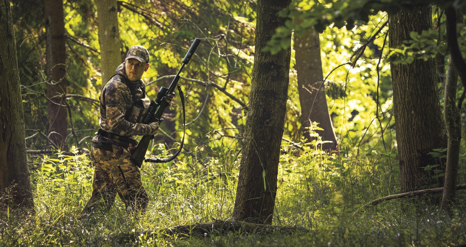 Stalker in lightweight camouflage layers carrying a moderated rifle through cover