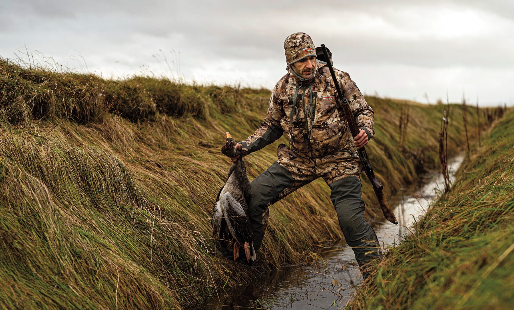 Wildfowler in Sitka camouflage clothing carrying a goose on the foreshore