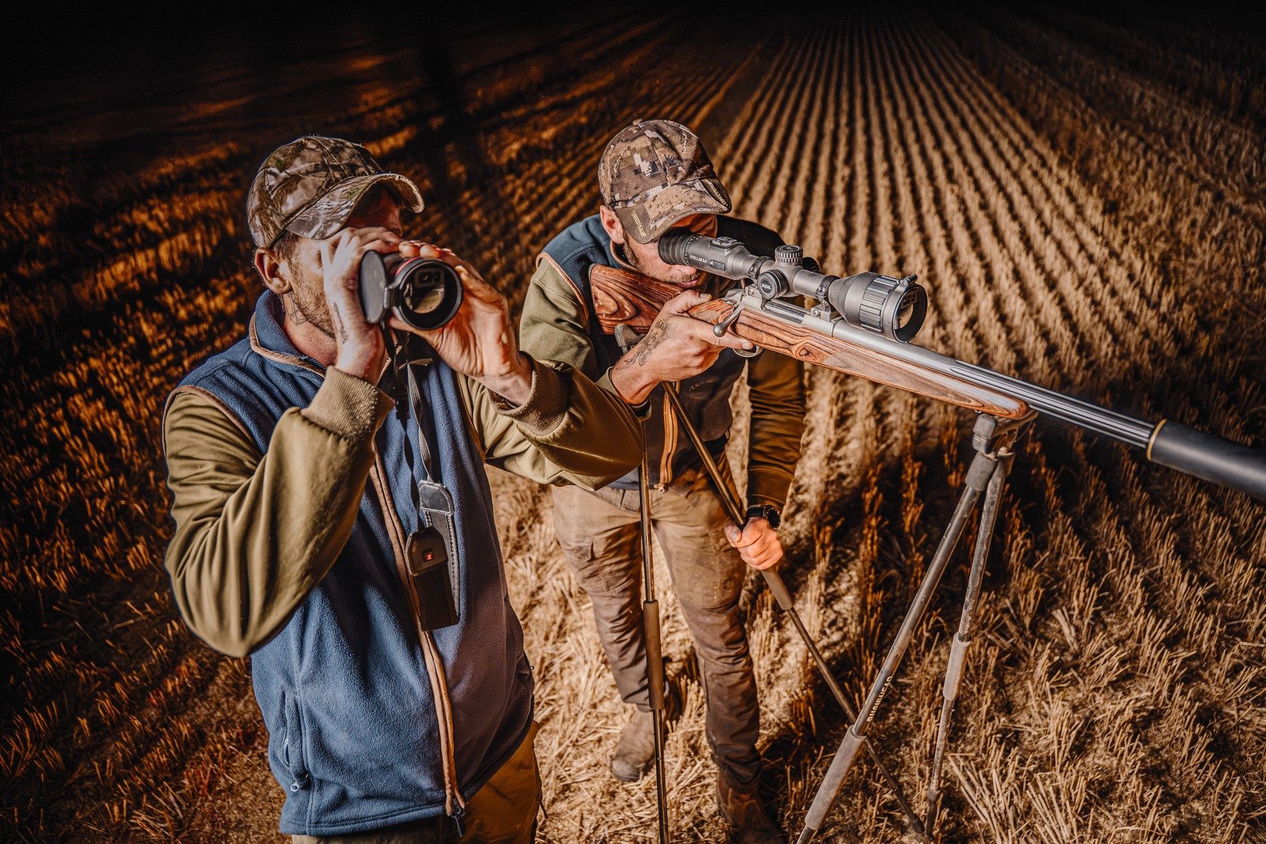 Spotter scanning with a thermal monocular while the rifleman waits on shooting sticks in a stubble field at night