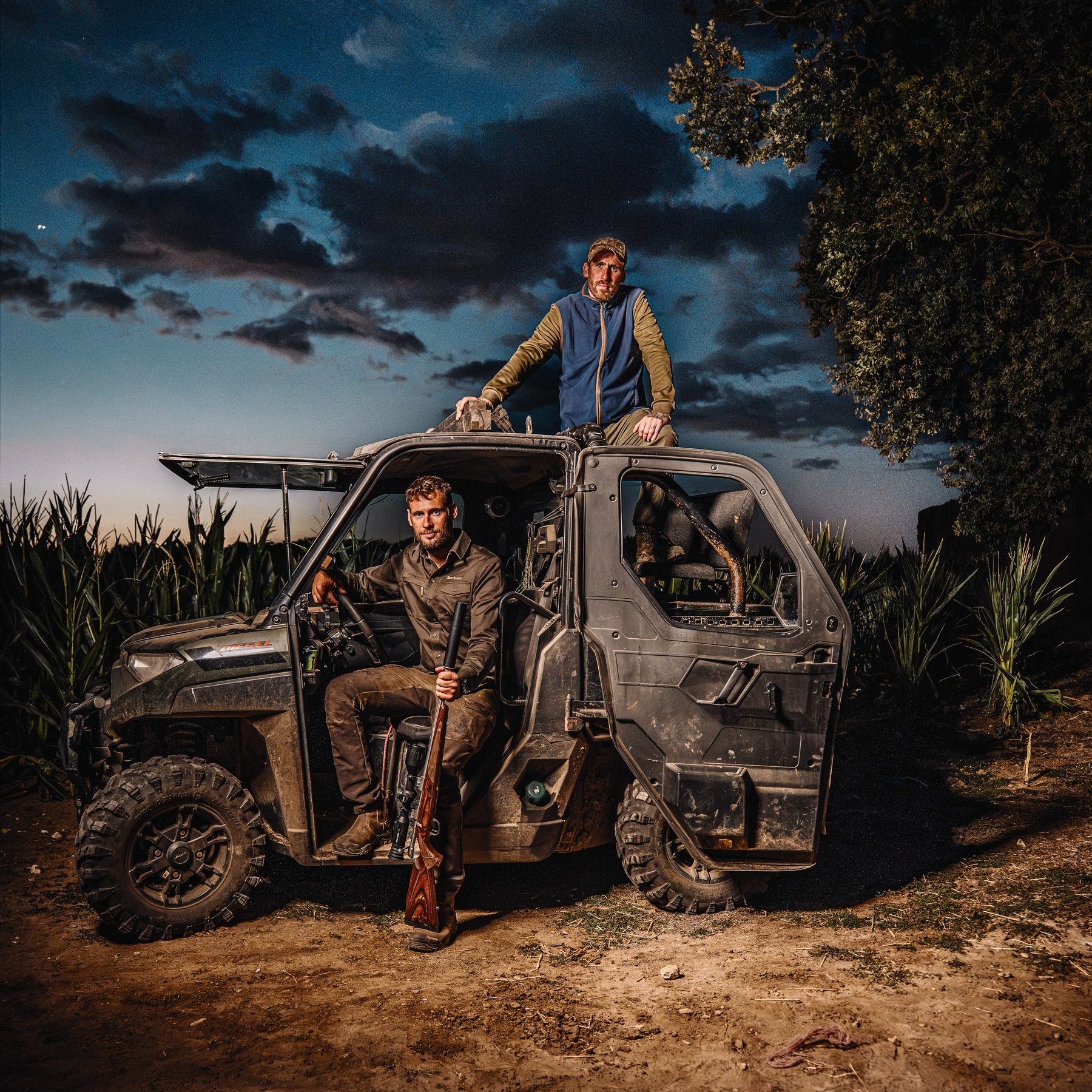 Two foxers with a rifle on a Polaris Ranger at dusk beside standing maize, ready for a night of fox control
