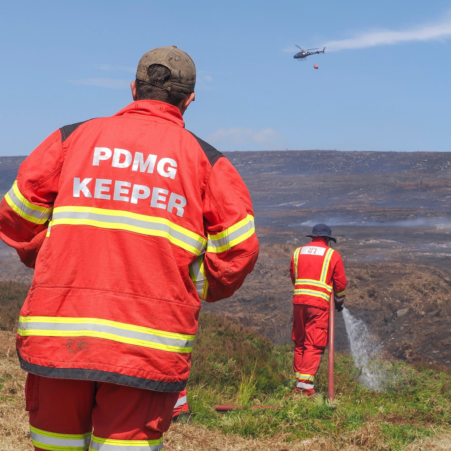 Peak District Moorland Group keeper watches a water-bucket helicopter as a colleague damps down the wildfire