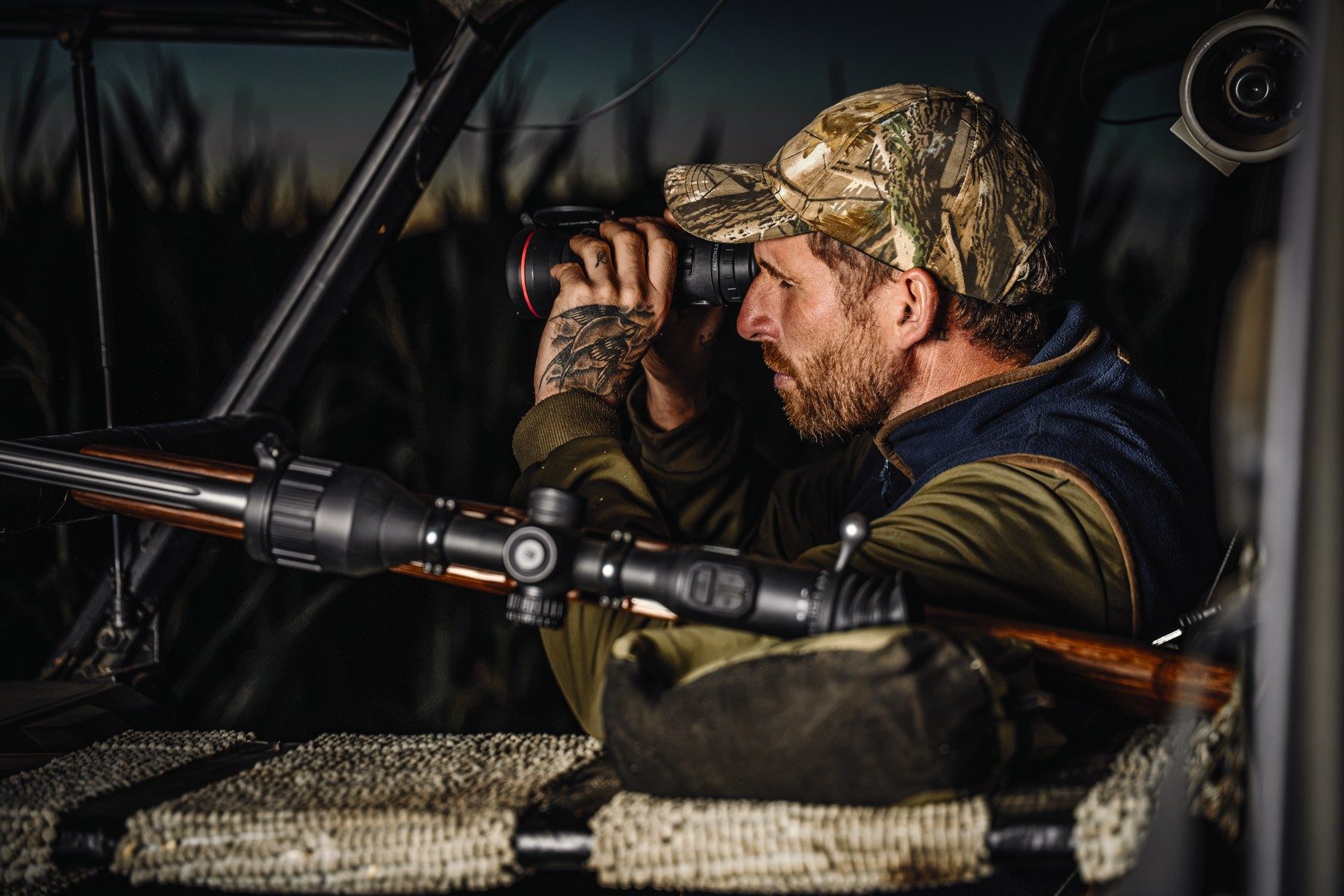 Spotter scanning stubble with a thermal monocular from the Polaris window, rifle resting on a carpeted support