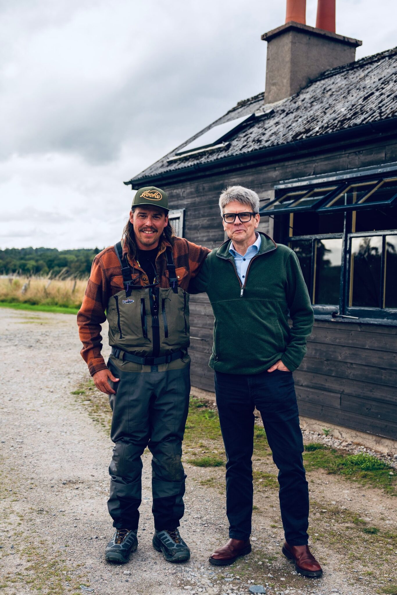 Angler in a fishing waistcoat and cap standing with a man in a green jumper outside a timber building